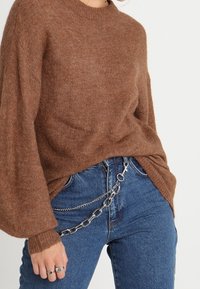 Hnědý pletený svetr s dlouhými, oversized rukávy v kombinaci s modrými, vysokým pasem džínami s stříbrným řetízkovým opaskem.