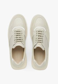 Světlé béžové tenisky vyrobené z hladké a texturované kůže. Obsahují perforovaný špičku, šněrování a gumovou podrážku s mírným vzorkem.