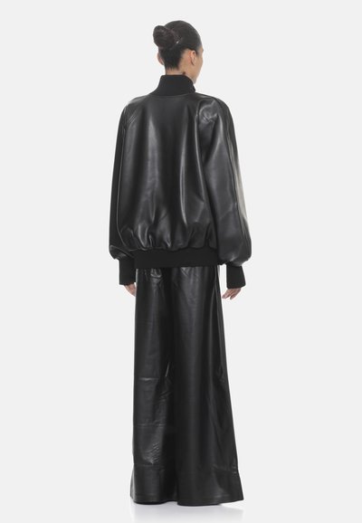 Giubbotto bomber in pelle nera con colletto e polsini a coste, abbinato a pantaloni in pelle nera a gamba larga. Texture liscia e vestibilità over.