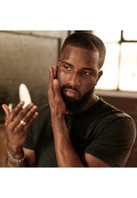 T-shirt noir sur une personne ayant les cheveux courts, affichant un teint de peau marron moyen. Présente un bracelet en chaîne tressée et une bague à motifs sur sa main gauche.