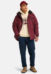 Maroon puffer jacket with black accents, beige hoodie featuring a graphic print, dark blue trousers, and tan hiking boots.
