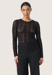 Žena v upnutém černém květinovém krajkovém topu s dlouhým rukávem a černých elegantních kalhotách, stojící na neutrálním světlém pozadí.