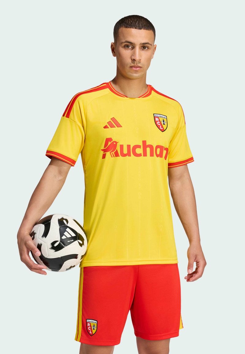 Jeune homme en tenue de football jaune et rouge tenant un ballon de football noir et blanc sur un fond clair uni.