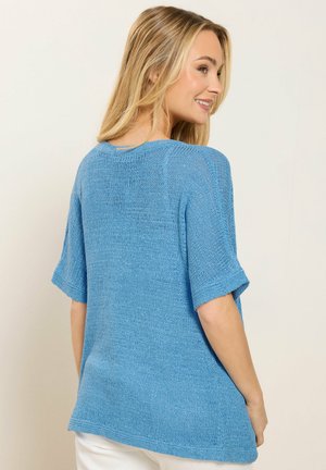 Nainen hymyilee ja katsoo sivulle, pukeutuneena väljästi istuvaan, lyhythihaiseen siniseen neulepaitaan ja valkoisiin housuihin yksiväristä vaaleaa taustaa vasten.