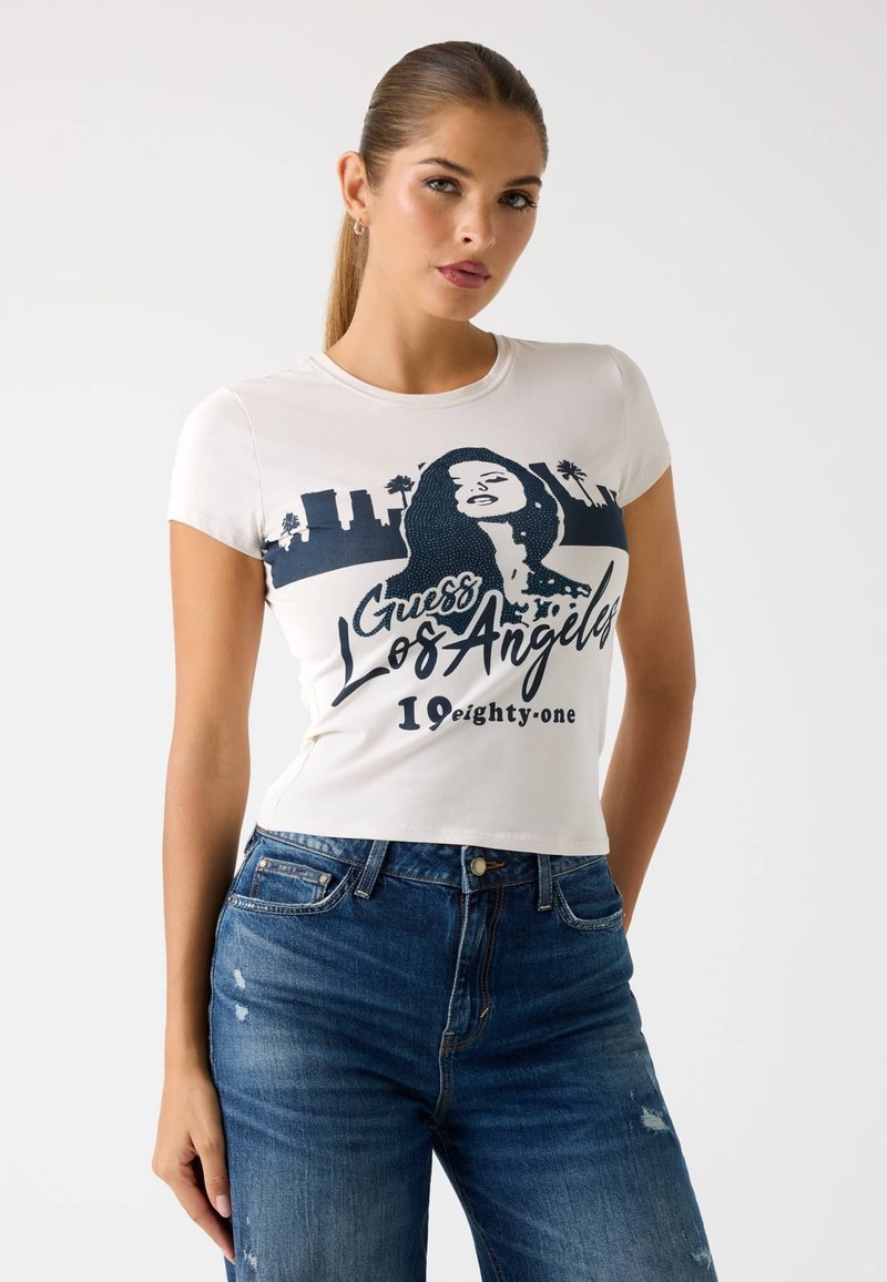 Biely skrátený tričko s tmavomodrou grafikou ženy a textom "Guess Los Angeles 19 eighty-one". Bavlna, krátke rukávy, priliehavý strih.