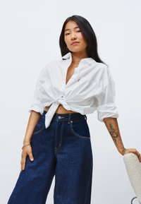 Chemise blanche nouée à manches longues ; jean large taille haute bleu foncé ; tatouage notable sur le bras ; fond uni.