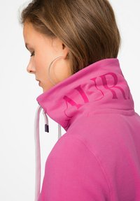 Felpa rosa con colletto alto, caratterizzata da una stampa discreta del marchio, chiusura con coulisse e una texture morbida e liscia.