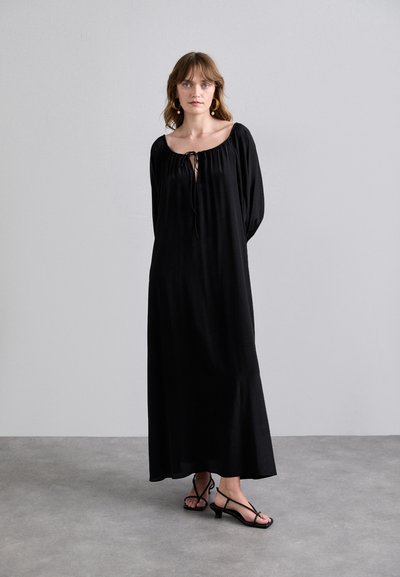 Black, long-sleeve maxi dress with gathered neckline and tie closure. Soft texture, flowy shape. Worn with black strappy sandals.
