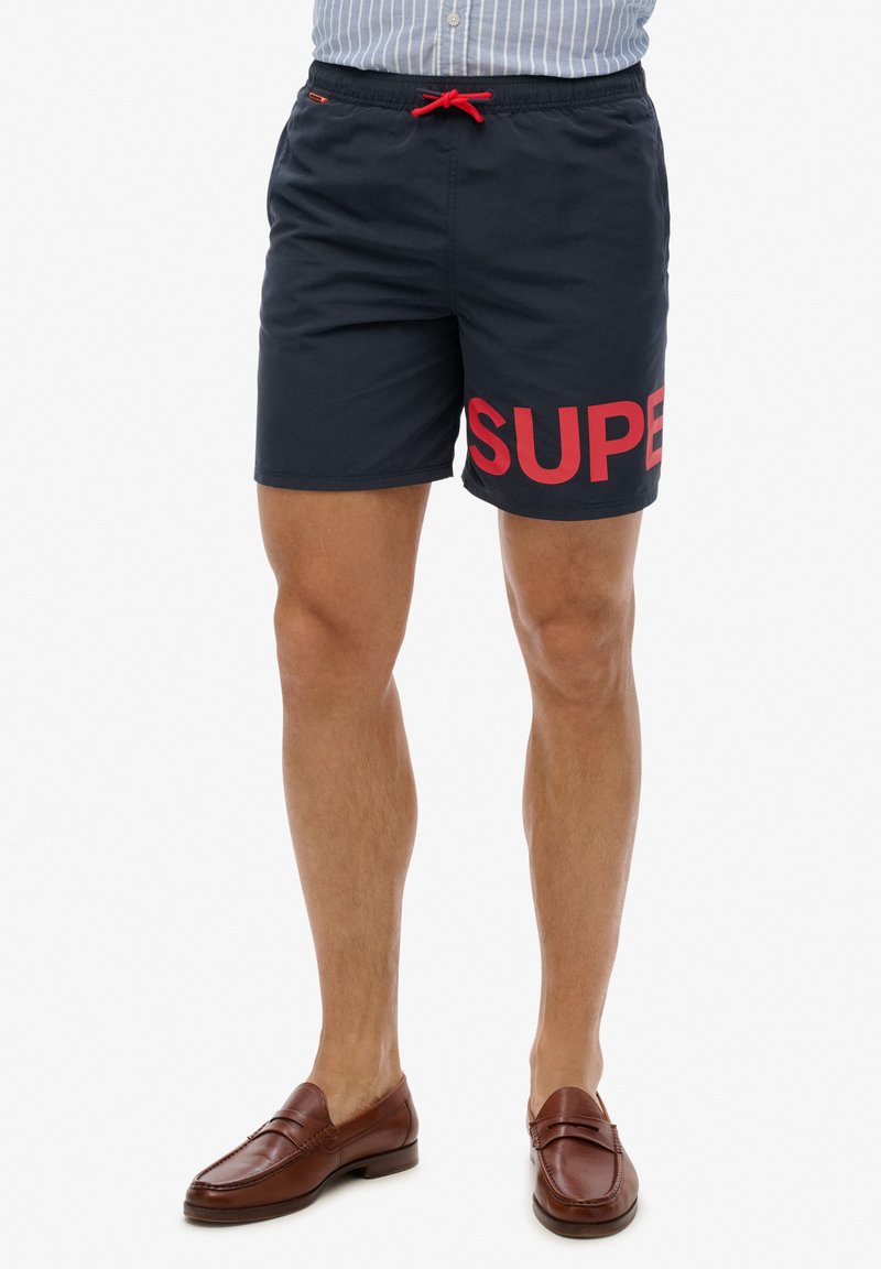 Shorts de baño de color azul marino hechos de material de secado rápido, con una destacada impresión roja de "SUPER" en la pierna izquierda y cintura con cordón.