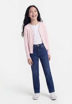 Κοπέλα που χαμογελά με μακριά σκούρα μαλλιά φοράει ανοιχτό ροζ ζακέτα, λευκό πουκάμισο, μπλε τζιν και λευκά αθλητικά παπούτσια, στέκεται σε λευκό φόντο.