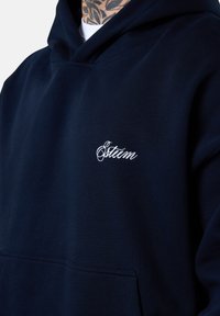 Sweatshirt à capuche bleu marine en tissu mélangé coton. Comprend une poche frontale et un logo brodé blanc sur la poitrine. Texture lisse.