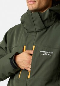 Grön utomhusjacka med gula dragkedjor, logotyp på bröstet, hög krage och justerbara ärmslut. Tillverkad av slitstarkt, strukturerat material.