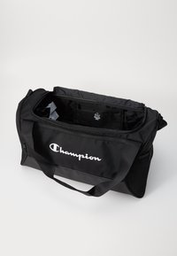 Black duffel bag made of durable fabric, featuring a zippered main compartment, side mesh pocket, and white "Champion" logo on the front.