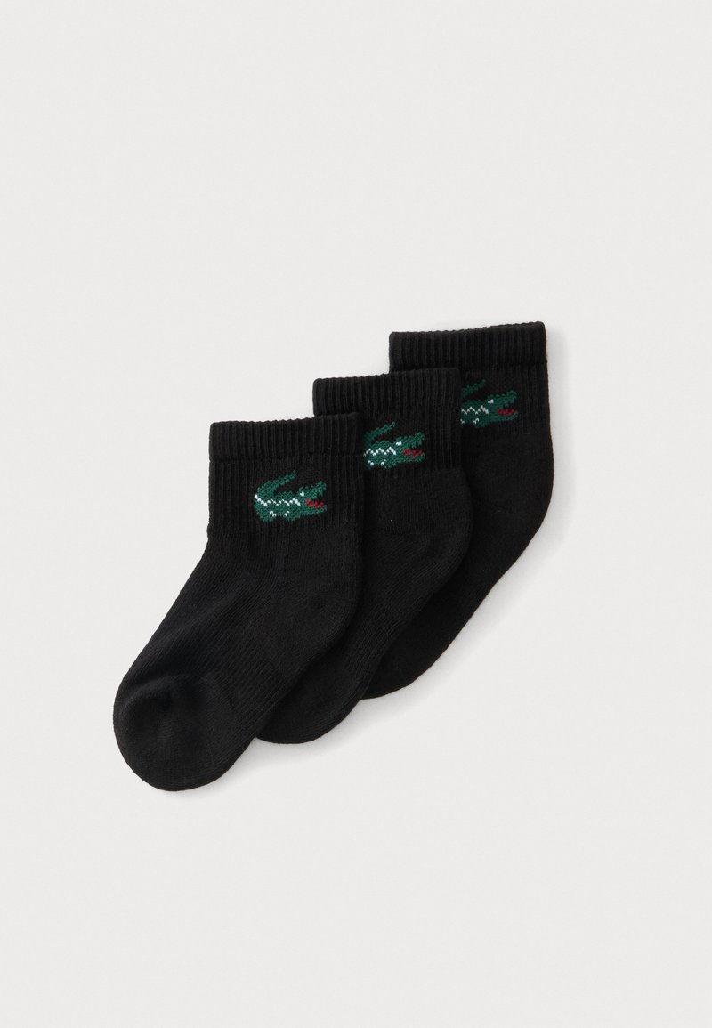 Trois chaussettes noires chevilles avec des logos de crocodile verts disposées en rangée légèrement superposée sur un fond blanc.