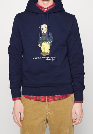 Uomo con felpa con cappuccio blu navy con grafica Polo Bear sotto una giacca, maglione a righe, pantaloni gialli, abbinati a camicia a quadri rossa e pantaloni in velluto a coste color sabbia.