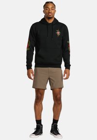 Zwarte hoodie met een voorkeurszak, groene slangafbeelding op de borst en rode accenten op de mouwen, gecombineerd met beige shorts en zwarte sneakers.