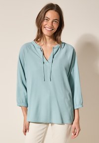 Top azzurro chiaro, con texture, dotato di scollatura a V e dettagli con lacci. Presenta maniche a tre quarti, vestibilità comoda e tessuto morbido.