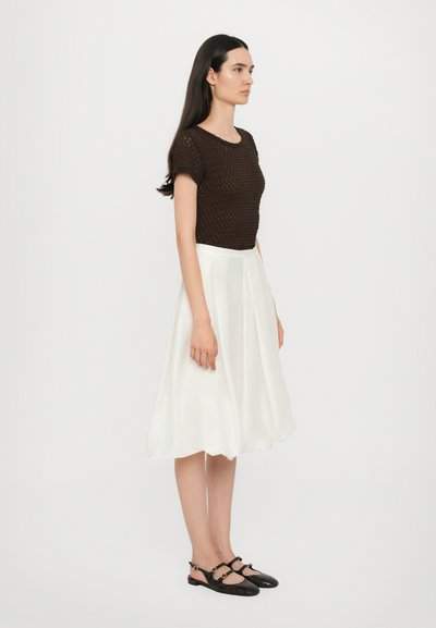 Bruine gebreide top met korte mouwen, witte midirok met plooien en zwarte slingback schoenen met gouden accenten. Effen achtergrond.