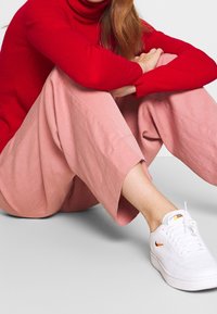 Röd polotröja, rosa vida byxor och vita sneakers med orange detaljer. Mjuka material och lösa passformer, sittande mot en neutral bakgrund.