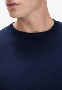 Mörkblå stickad tröja med rund halsringning, med en mjuk textur och ribbad kant. Närbild av kragen och nackområdet.