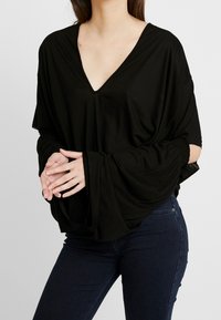 Haut noir ample avec un décolleté en V profond et des manches froncées, confectionné dans un matériau doux. Associé à un jean foncé, mettant en avant un design décontracté.