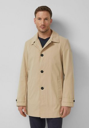 Man met een beige jas met zwarte knopen over een donker overhemd, staand tegen een effen lichtgrijze achtergrond.