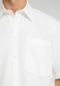 Camicia bianca a maniche corte realizzata in tessuto leggero, con colletto a punta, chiusura con bottoni e una singola tasca sul petto.