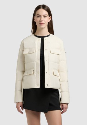 Femme portant une veste matelassée crème avec poches à rabat sur une robe noire, debout devant un fond gris clair uni.