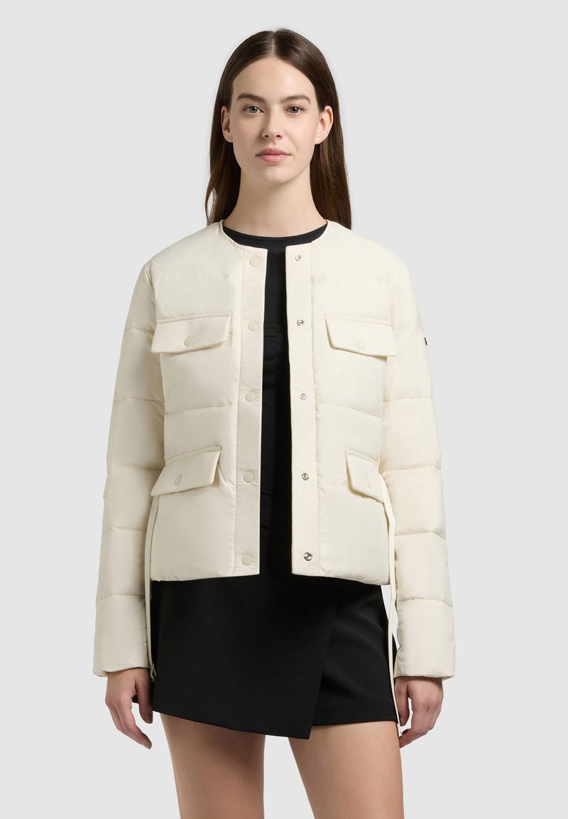 Donna che indossa una giacca trapuntata color crema con tasche a patta sopra un vestito nero, in piedi davanti a uno sfondo grigio chiaro uniforme.