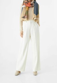 Femme portant un pantalon blanc à jambes larges, un haut beige, une écharpe colorée à motifs abstraits, et des bottines beiges à talons, debout les mains dans les poches.