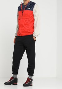 Veste coupe-vent rouge et marine avec une poche avant, associée à un pantalon de jogging noir et des bottes de randonnée noires avec des lacets rouges. Chaussettes grises visibles.