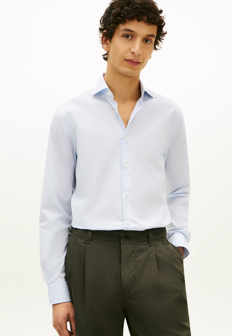 Jeune homme aux cheveux bouclés portant une chemise bleu clair boutonnée et un pantalon vert foncé, debout avec une main dans la poche.
