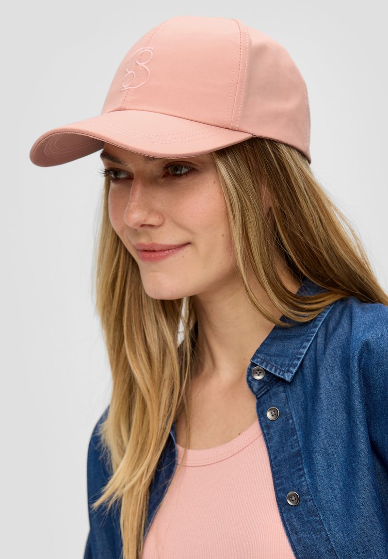 Roze baseballcap met gestikt logo, gebogen klep en gestructureerde stof. Draagt schuin met haar dat naar beneden valt.