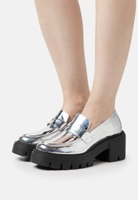 Des mocassins argentés métalliques avec une semelle de plateforme noire épaisse. Surface lisse et brillante avec des coutures détaillées et une double bride sur le devant.