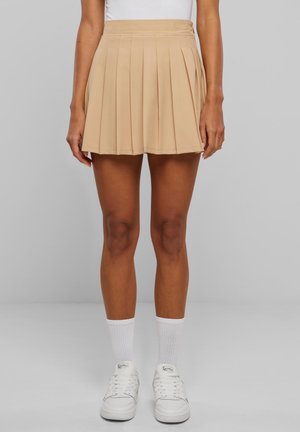 Femme portant une mini-jupe plissée beige, des baskets blanches et des chaussettes blanches montant jusqu'au mollet, debout devant un fond gris uni.