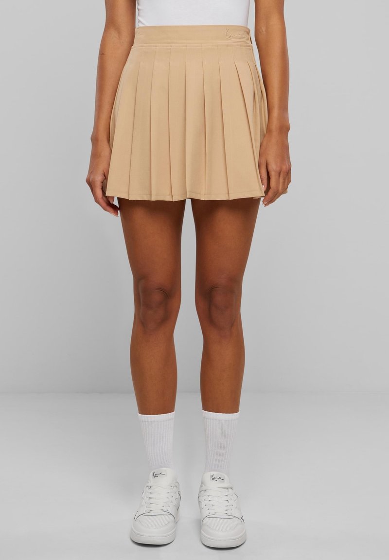 Femme portant une mini-jupe plissée beige, des baskets blanches et des chaussettes blanches montant jusqu'au mollet, debout devant un fond gris uni.