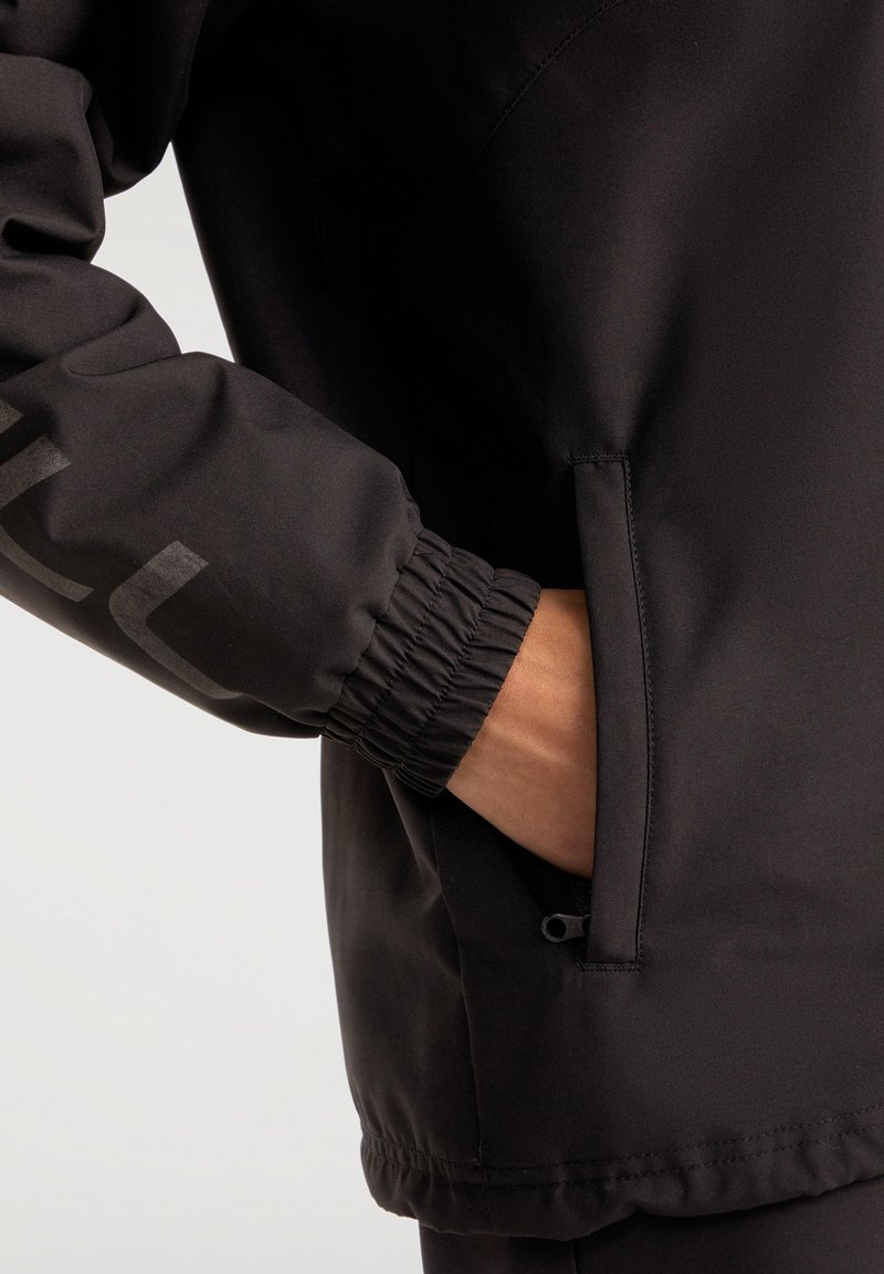 Chaqueta negra con tejido texturizado, puños elásticos y un bolsillo lateral con cremallera. El diseño es minimalista sin logotipos ni patrones visibles.