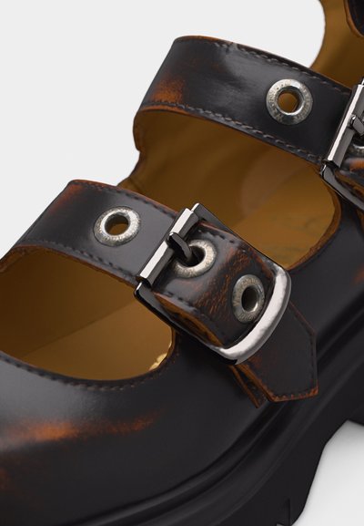 Κοντινό πλάνο σκούρου καφέ δερμάτινου παπουτσιού με παχιά σόλα, δύο λουριά με ασημένιες αγκράφες και λεπτομέρειες με οπές.