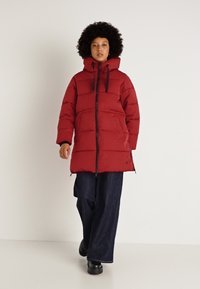 Rød pufferjakke med hætte, der har en frontlynlås og snoretræk, sammen med løstsiddende mørke denimbukser og sorte sko.