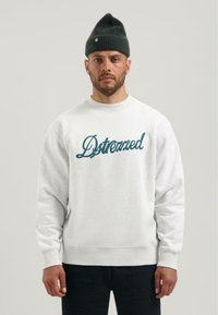 Felpa grigia chiaro con colletto a girocollo, scritta ricamata blu, polsini e orlo a coste. I pantaloni neri e un berretto verde scuro completano l'outfit.