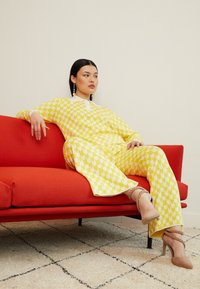 Żółty wzorzysty strój z motywami serc i białym kołnierzykiem, w połączeniu z beżowymi sandałami na obcasie. Modelka siedzi na czerwonej sofie przed beżową ścianą.