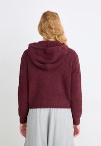 Bordo skrajšan hoodie iz mehkega, teksturiranega materiala z vrvico za nastavljanje kapuce in rebrastim robom, v kombinaciji s svetlo sivo širokimi hlačami.