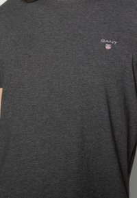 Mørk grå bomulds T-shirt med korte ærmer, der har et lille hvidt "GANT"-logo og et skjold emblemet med røde accenter på den øverste venstre bryst.