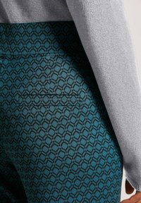 Türkisfarbene Hosen mit einem geometrischen Muster in Schwarz, einer Gesäßtasche und einem teilweise sichtbaren grauen Pullover.