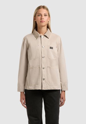 Jauna moteris dėvi smėlio spalvos užsegamą švarką su didelėmis priekinėmis kišenėmis ir juodas kelnes, stovinti priešais paprastą šviesų foną.