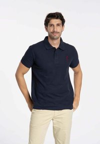 Polo-shirt bleu marine avec col et patte de boutonnage à trois boutons, arborant un logo rouge sur la poitrine gauche. Porté avec un pantalon beige.