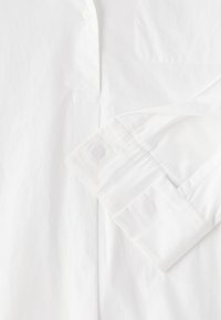 Camicia bianca con bottoni, caratterizzata da una superficie liscia, dettagli con polsini piegati e una tasca sul petto. Design pulito con accenti minimi.