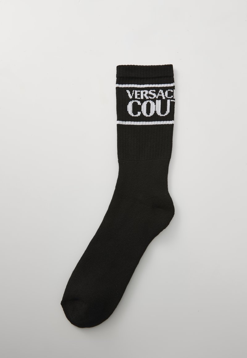 Črne telečje nogavice z rebrastim robom, ki vsebujejo bel napis "VERSACE COUTURE" in vodoravne bele črte na svetli podlagi.