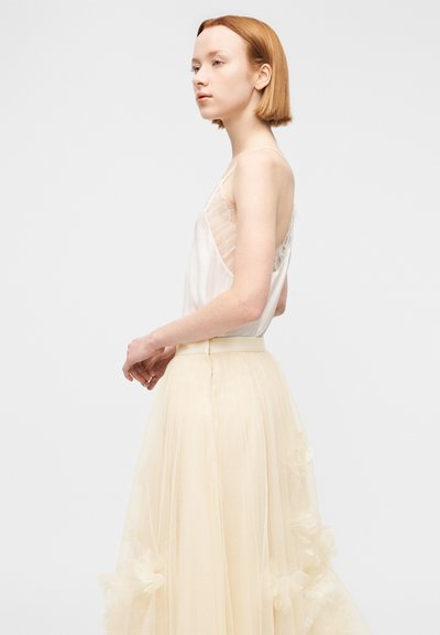 Νεαρή γυναίκα με κοντά κόκκινα μαλλιά, φοράει λευκό αμάνικο τοπ και κρεμ τούλινη φούστα με λουλουδάτες λεπτομέρειες, σε ποζέ προφίλ.