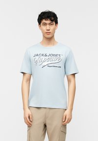 Νεαρός με κοντά σγουρά μαλλιά που φοράει ένα ανοιχτό μπλε T-shirt "Jack & Jones Superior" και μπεζ παντελόνι cargo σε λευκό φόντο.